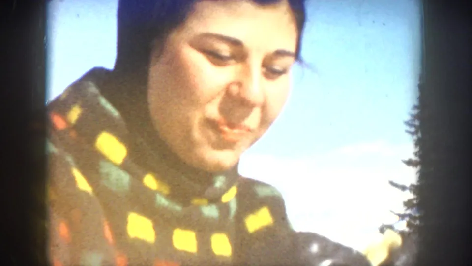 1961 Aspen home-movie frame of a woman smiling in a colorful scarf, with bright winter sky, snow, and evergreens nearby.