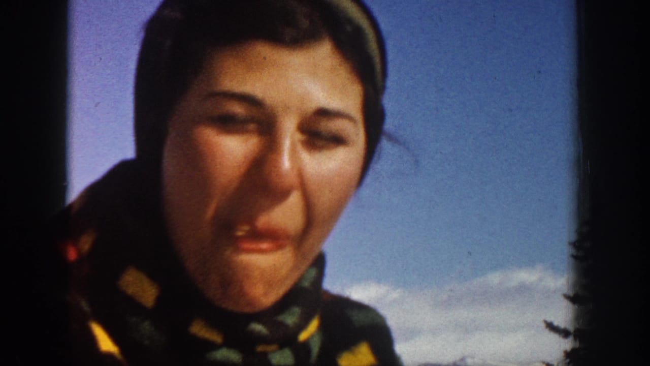 A woman in winter clothing faces the camera in 1961 Aspen, with snow, blue sky, and pine trees visible behind her.