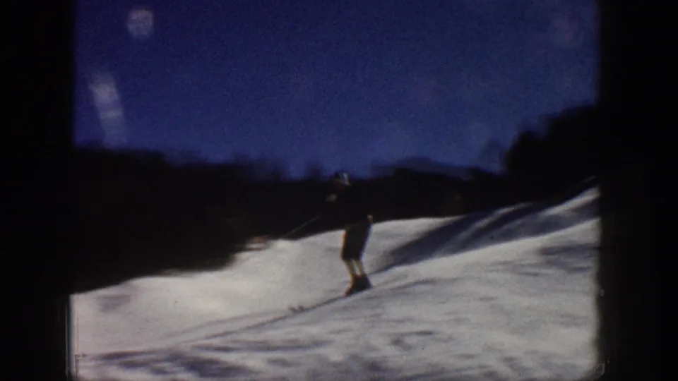 Aspen, Colorado 1961 frame with a distant skier using poles on a snowy slope beneath dark tree and mountain silhouettes.