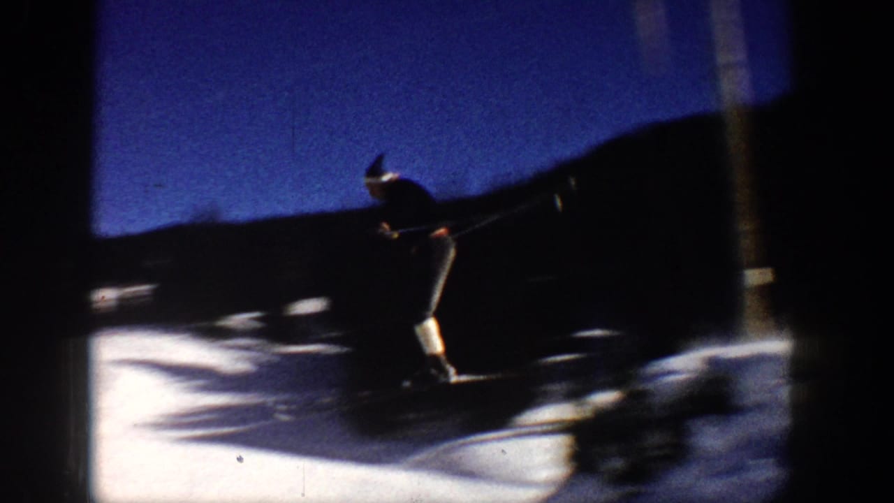 Skier in dark jacket and light pants moving across a snowy slope near dark tree shadows.