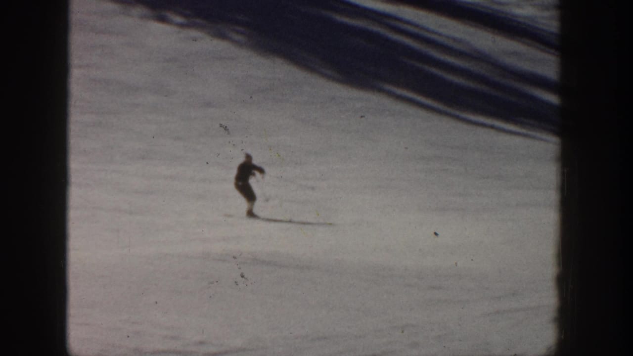 1961 Aspen home-movie frame showing a distant skier in dark clothing moving across a snowy ski slope.