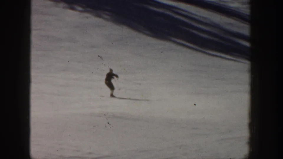 1961 Aspen home-movie frame showing a distant skier in dark clothing moving across a snowy ski slope.