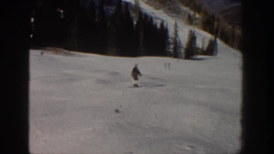 1961 Aspen home-movie frame showing the skier lower on the run with evergreens and mountain slope behind.