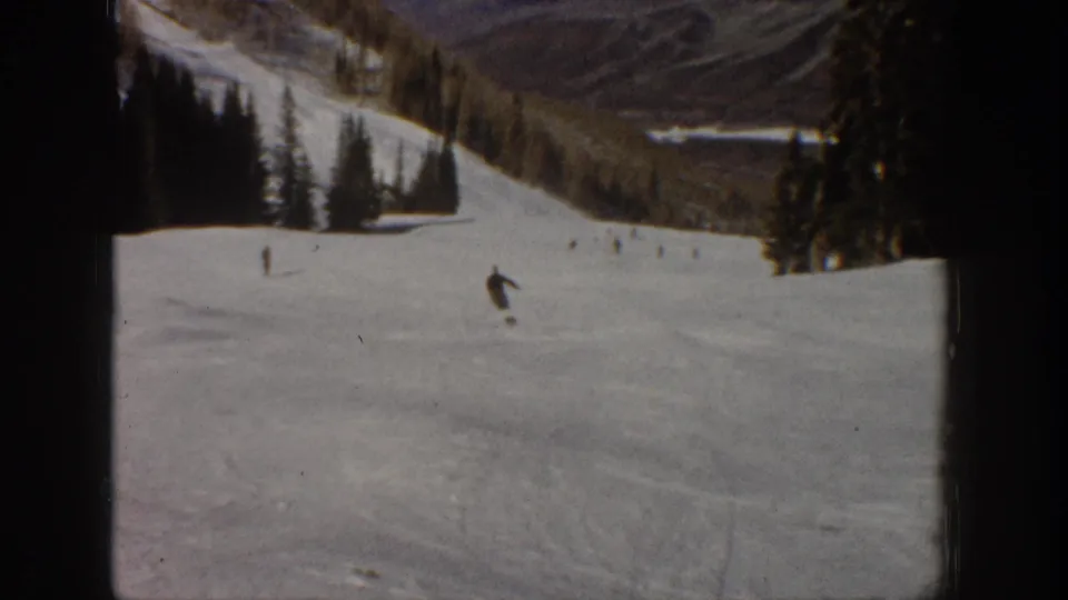 1961 Aspen ski slope frame showing a small skier descending toward the lower right with trees and mountains beyond.