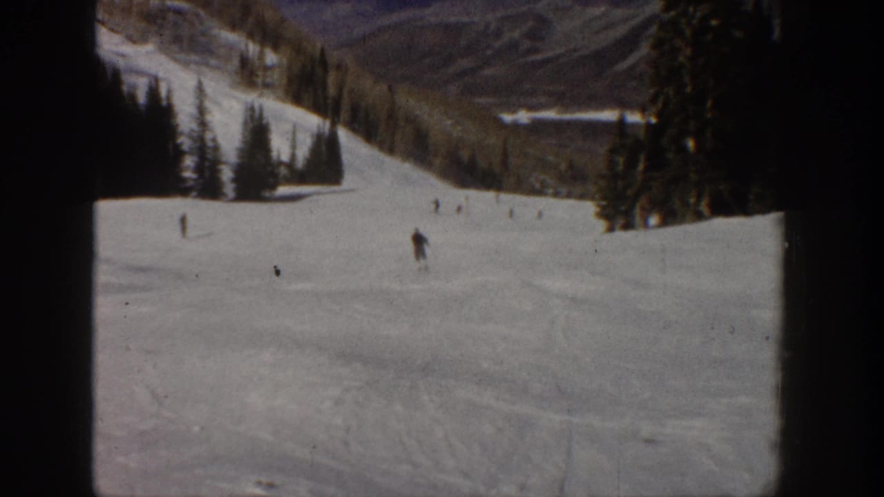 Aspen, Colorado 1961 home-movie frame showing small skiers descending a wide snowy slope with evergreens and mountain terrain beyond.
