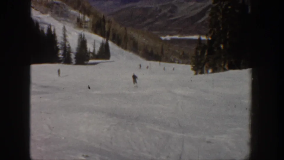 Aspen, Colorado 1961 home-movie frame showing small skiers descending a wide snowy slope with evergreens and mountain terrain beyond.