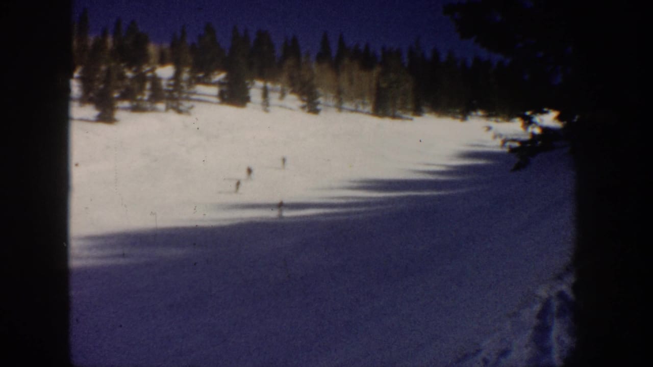 Aspen, Colorado 1961 frame with small skiers spread across a bright snowfield below evergreen trees and a clear blue sky.