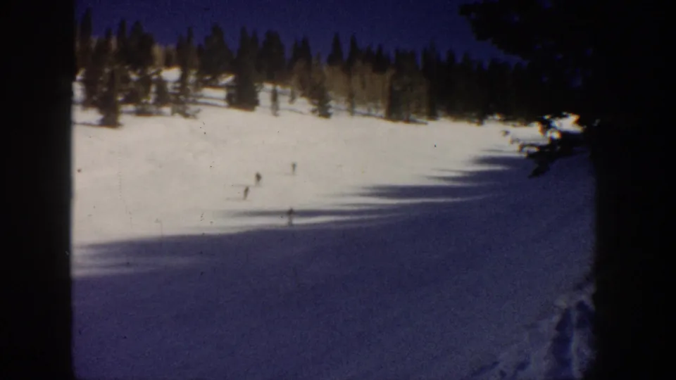 Aspen, Colorado 1961 frame with small skiers spread across a bright snowfield below evergreen trees and a clear blue sky.