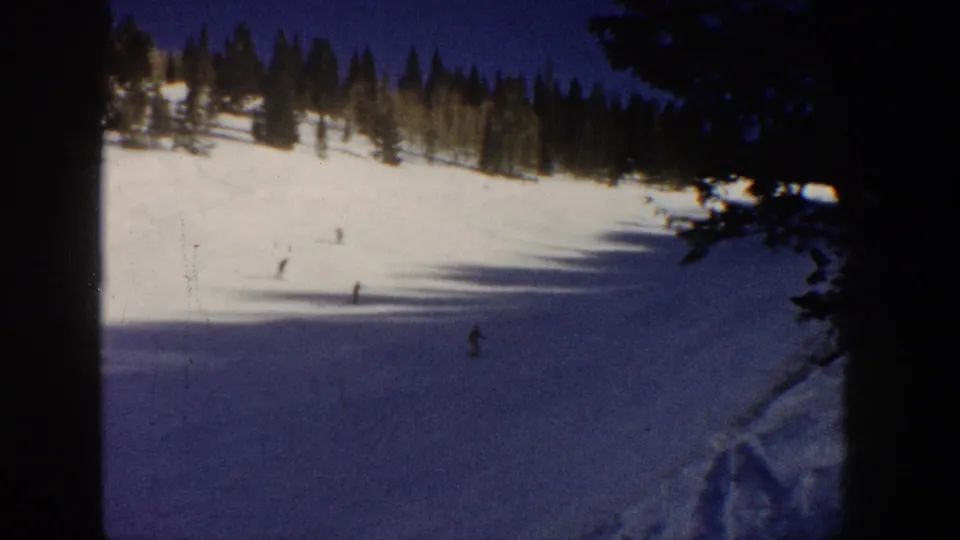 Aspen, Colorado 1961 home-movie frame showing skiers crossing a partly shadowed slope beneath a dark evergreen ridge.