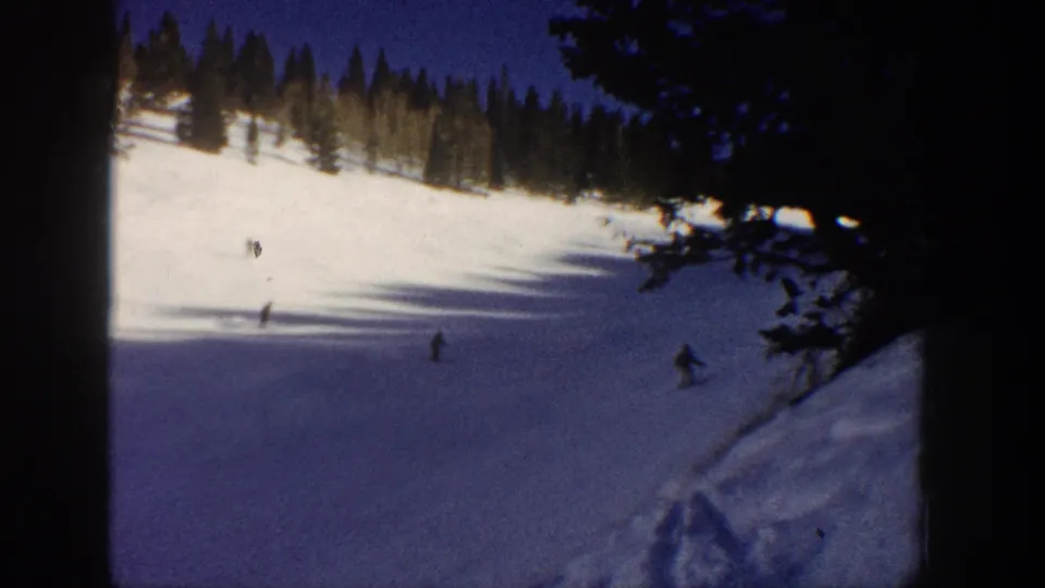 Aspen, Colorado 1961 frame showing multiple distant skiers on a snowy run, with evergreen trees and dark foreground branches at the right edge.