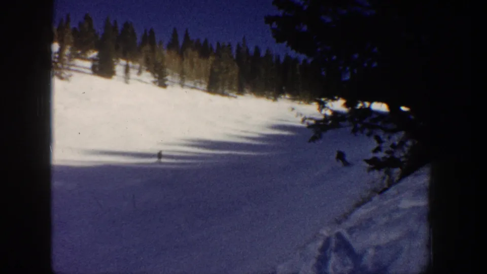 1961 Aspen 8mm frame showing distant skiers on a shaded ski slope with dark pines in the foreground.