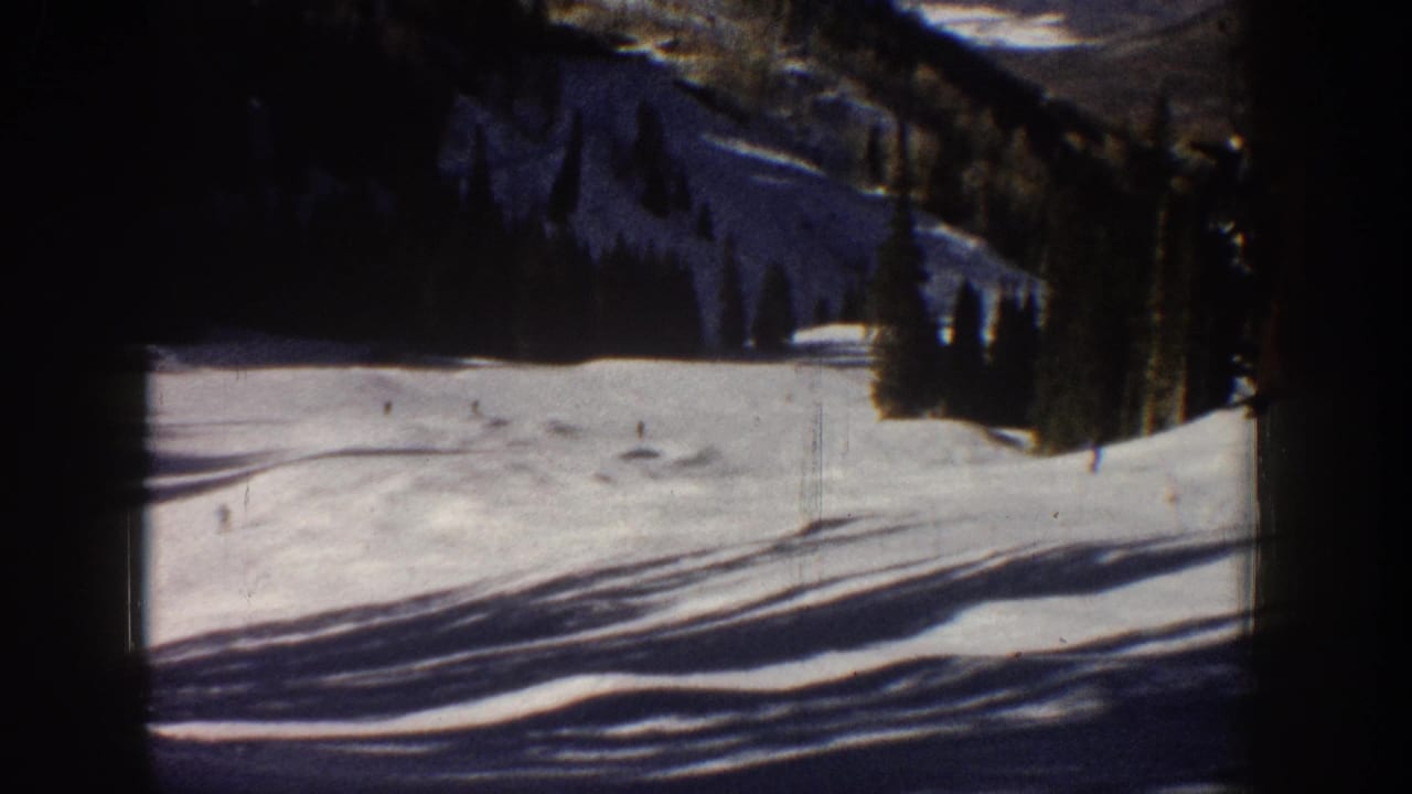 1961 Aspen home-movie frame showing tiny skiers on a bright snowy slope with evergreens and mountain terrain behind them.