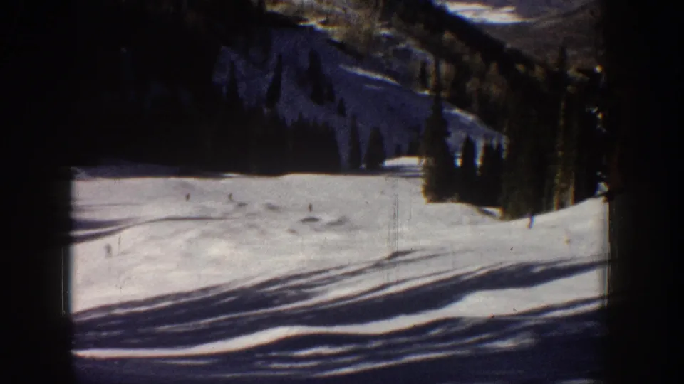 1961 Aspen home-movie frame showing tiny skiers on a bright snowy slope with evergreens and mountain terrain behind them.