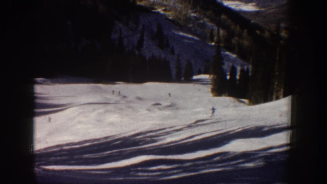 1961 Aspen 8mm frame with distant skiers on a sunlit snowfield, dark tree line, and rugged mountain slope beyond.
