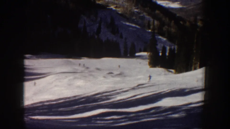 1961 Aspen 8mm frame with distant skiers on a sunlit snowfield, dark tree line, and rugged mountain slope beyond.