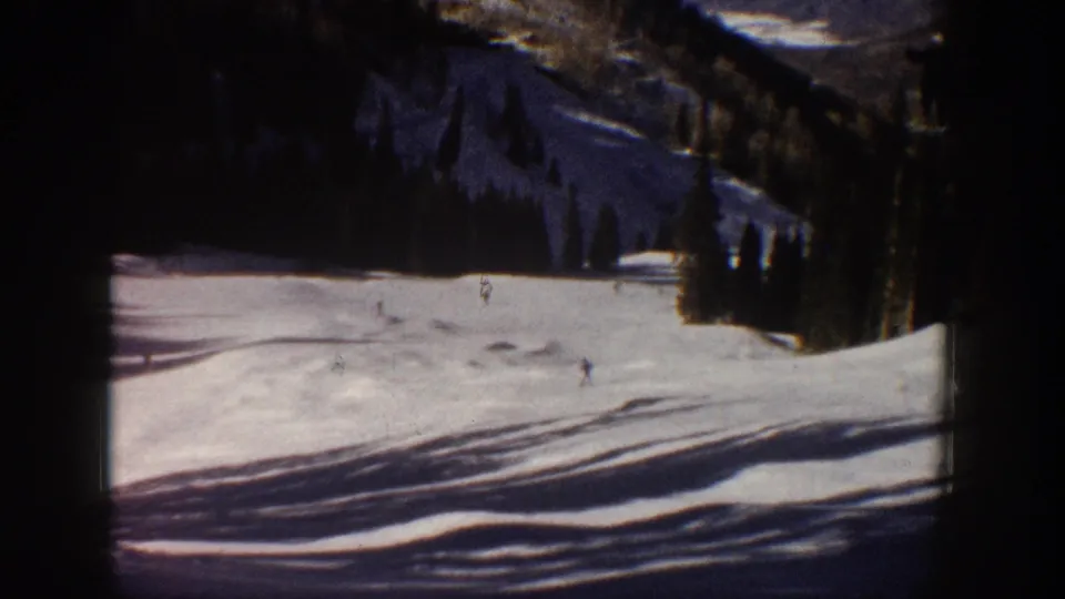 Final 1961 Aspen frame showing tiny skiers on a snowy run, backed by dark evergreens, mountain terrain, and visible 8mm film texture.