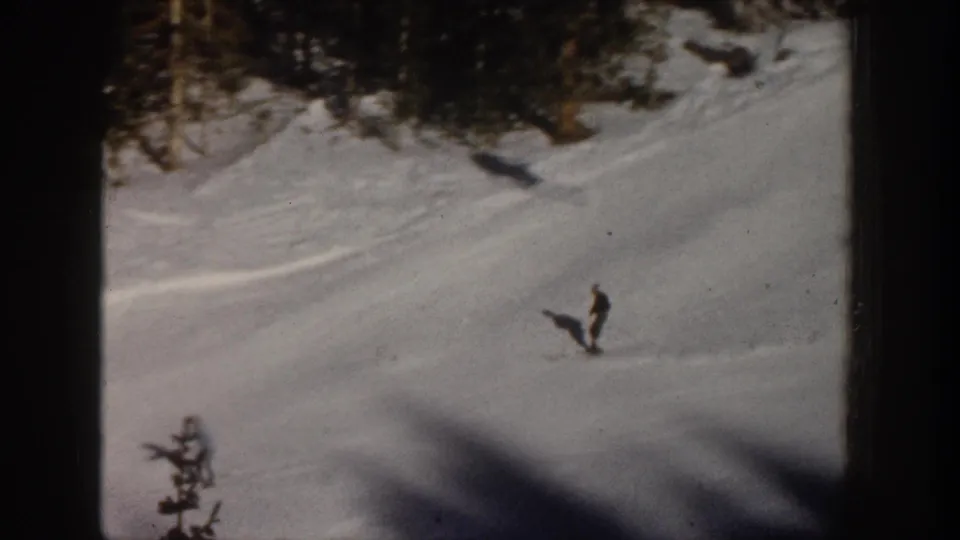 1961 Aspen home-movie frame showing a distant skier on open snow, with evergreens above and dark shadows across the lower frame.