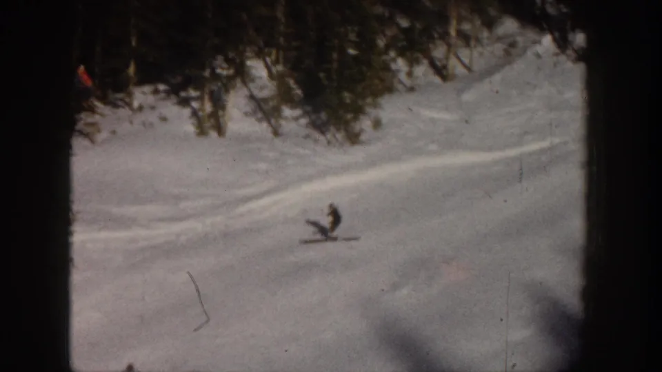 1961 Aspen frame showing a lone skier moving across a pale snowfield below a line of evergreen trees.