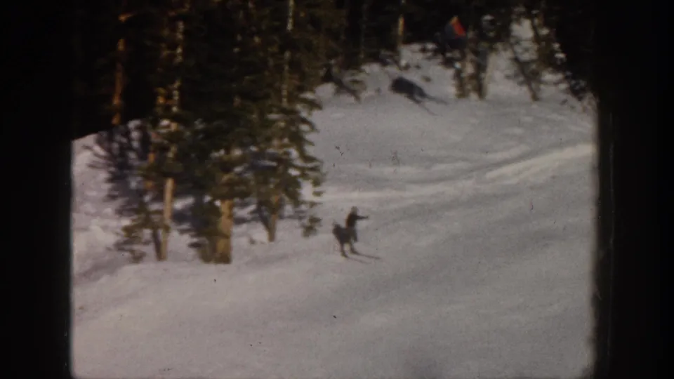 1961 Aspen 8mm frame of a skier descending a snowy run bordered by evergreens and faint tracks.