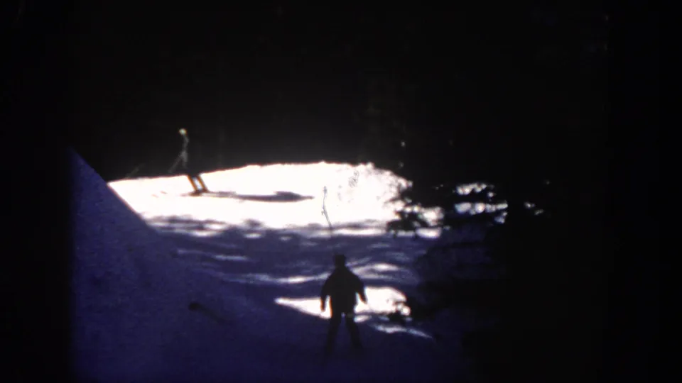 1961 Aspen home-movie frame with skier silhouettes crossing bright snow beside a shadowed forest edge.