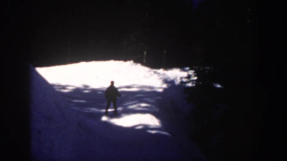 1961 Aspen 8mm frame showing a lone skier silhouette on sunlit snow below a dark wooded slope.