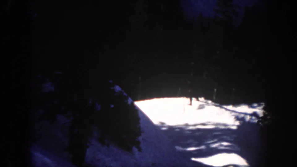 Night skiing continues on a bright snow path bordered by dark pines in the 1961 Aspen footage.