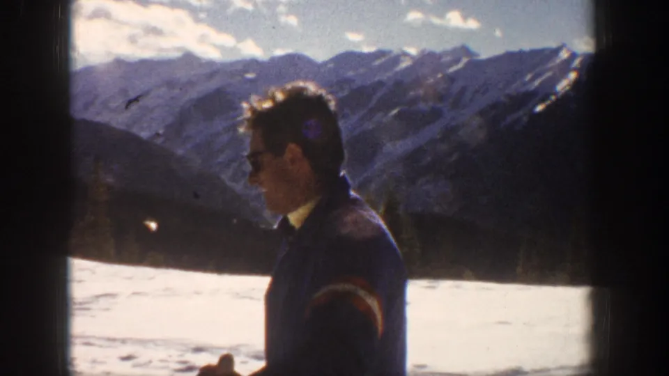 Adult man in sunglasses and a ski jacket stands in profile before snowy Aspen mountain ridges in 1961 footage.