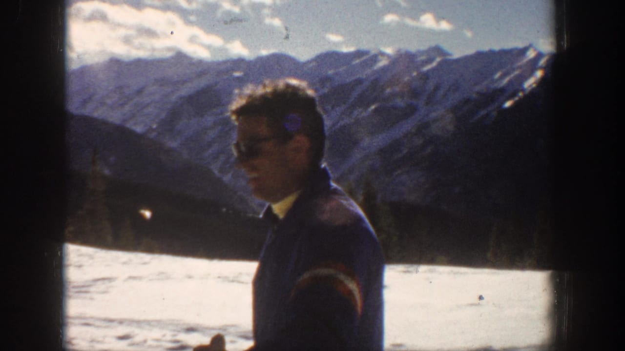 Adult man in a blue ski jacket appears in Aspen snow with dark mountain ridges behind him, filmed on 8mm home-movie stock.