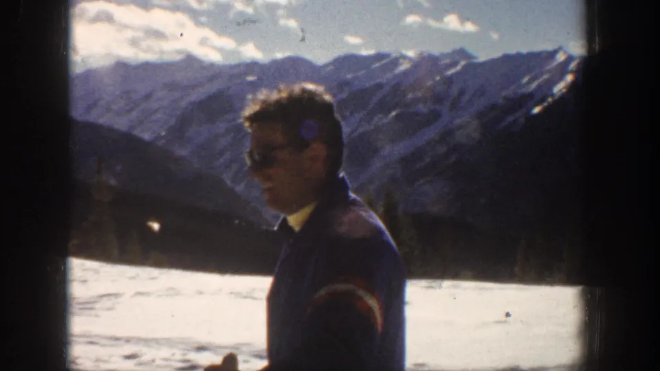 Adult man in a blue ski jacket appears in Aspen snow with dark mountain ridges behind him, filmed on 8mm home-movie stock.