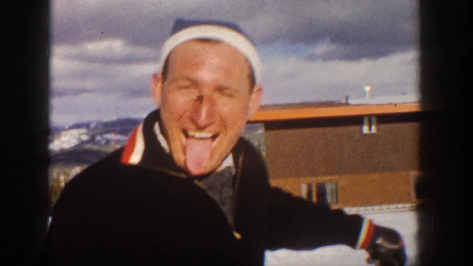 Adult man wearing a white headband and dark ski jacket jokes for the camera near snow, a low building, and distant mountains.