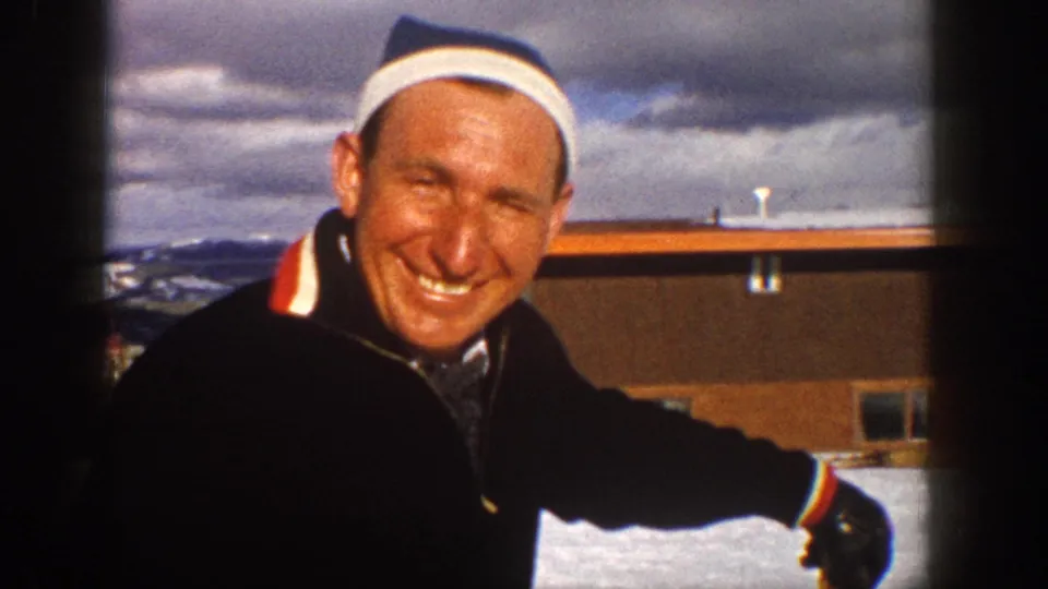 Smiling adult man in a white headband and dark ski jacket leans at an Aspen ski area, with snow, mountains, and a low building behind him.