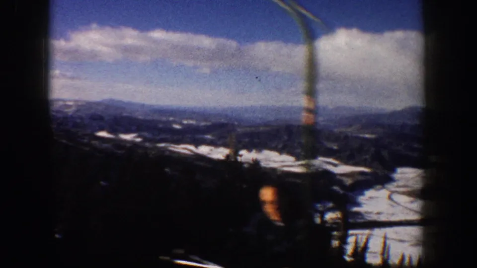 A handheld 8mm view of the Aspen mountain landscape shows snow patches, distant ridges, cloudy sky, and a partly visible person near the bottom edge.