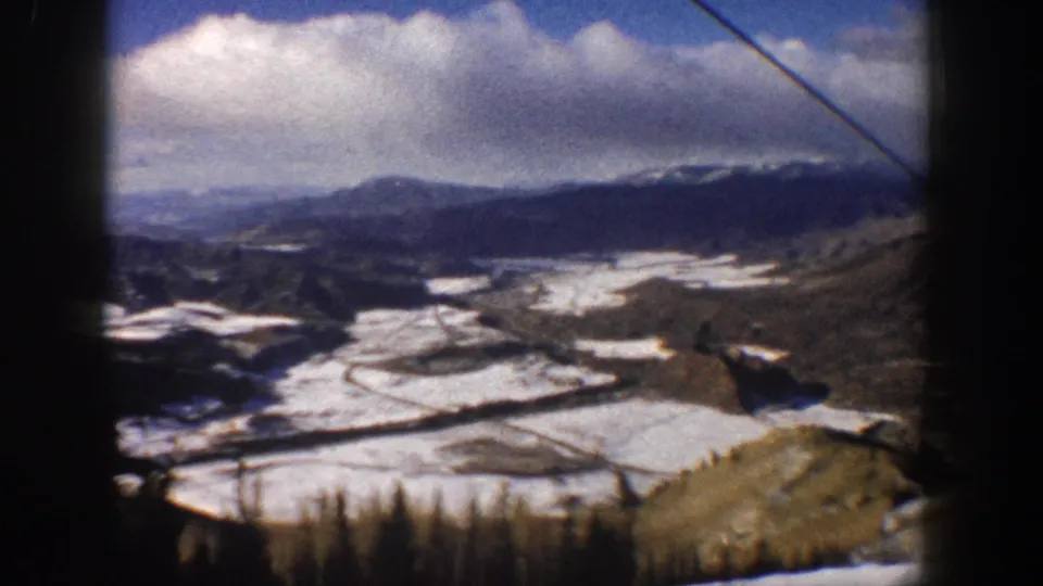 1961 Aspen lift descent frame showing a snow-streaked valley, conifers, and a dark mountain ridge beneath clouds.