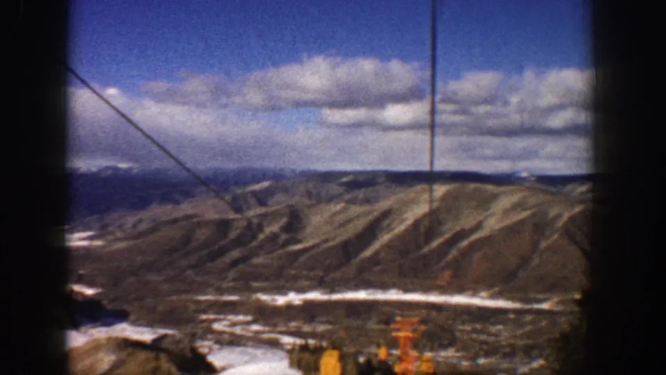 Visitor-shot 1961 Aspen ski-lift footage looking across a broad brown ridge, with snow patches, cable lines, and cloudy blue sky.