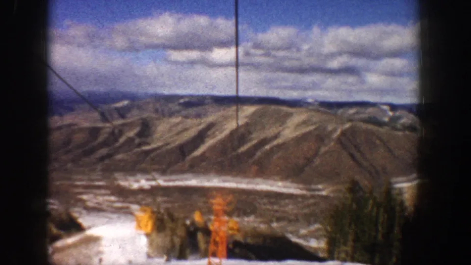 1961 Aspen lift view with yellow and orange lift hardware near a snowy slope edge, trees, and ridged mountains in the distance.