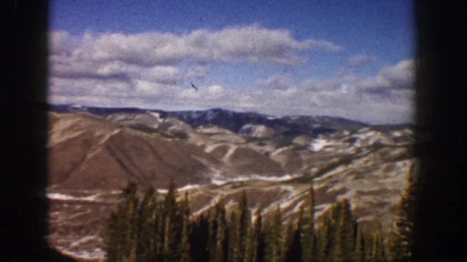 1961 Aspen-area 8mm frame with rolling Colorado mountain terrain, snow traces, evergreen tops, and a bright clouded sky.