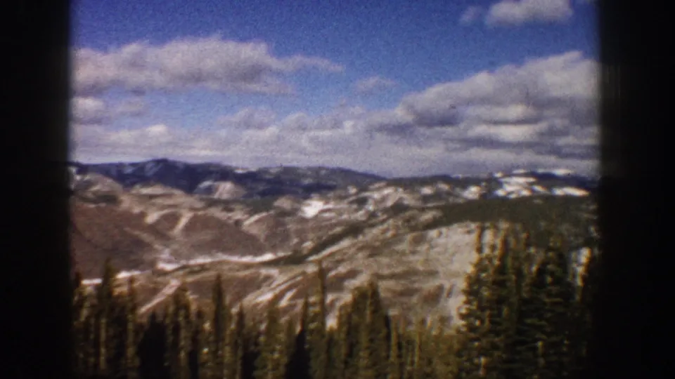1961 Aspen, Colorado 8mm view of evergreen forest foreground, pale mountain slopes, scattered snow, and a clouded blue sky.