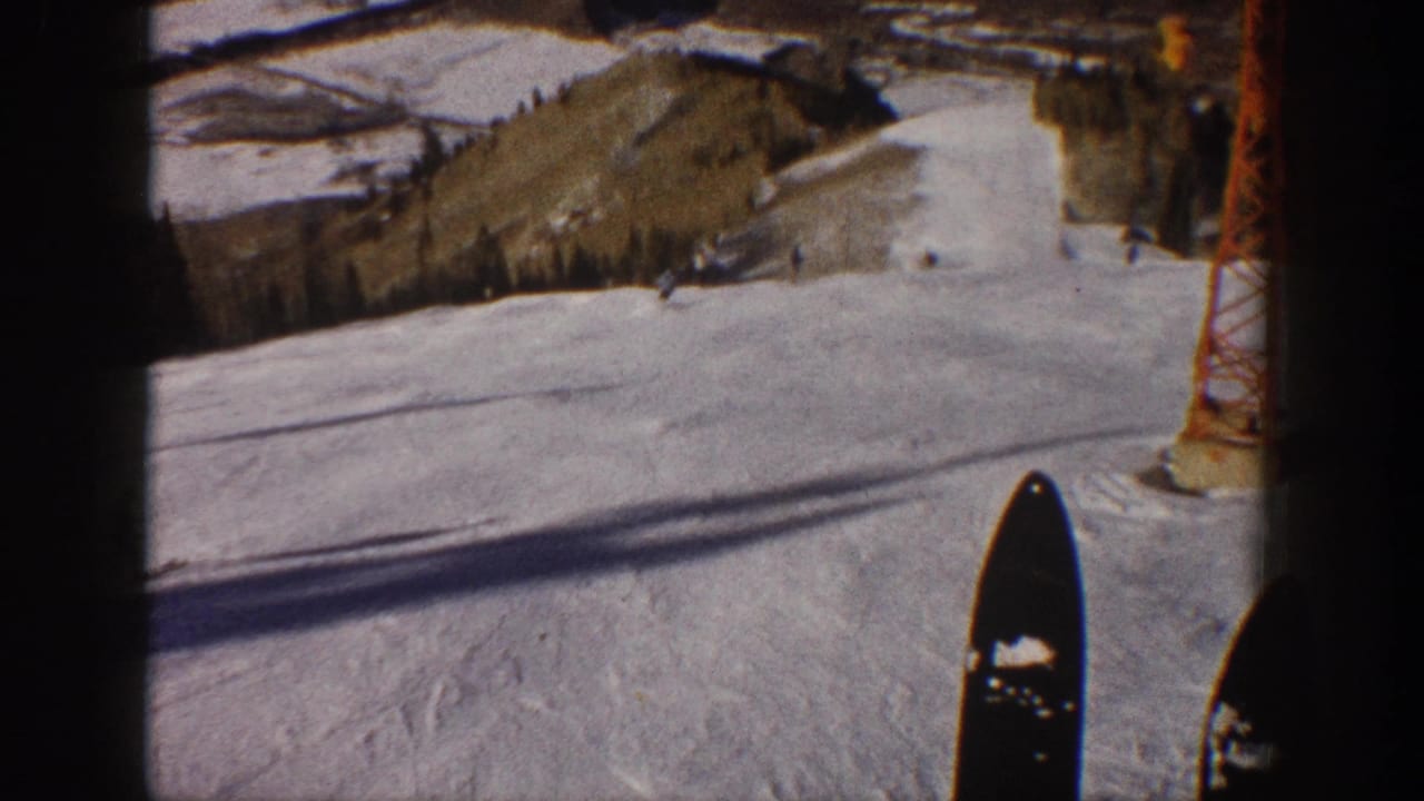 First-person-style Aspen ski-slope view with ski tips, a lift tower, snowy terrain, and small skiers below.