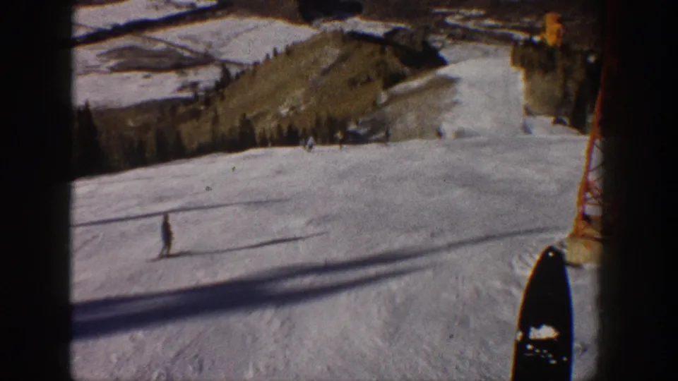Aspen ski run with a distant skier crossing the snow, a lift tower at right, and wooded terrain beyond.