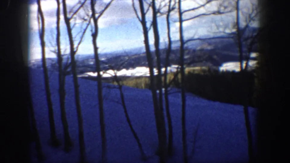 1961 Aspen, Colorado frame looking through bare aspen trunks toward a blue snow slope and distant winter hills.
