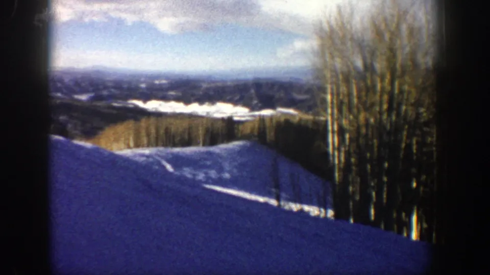 1961 Aspen, Colorado home-movie frame showing a snowy slope, bare aspen trees, distant mountains, and blue winter shadows.