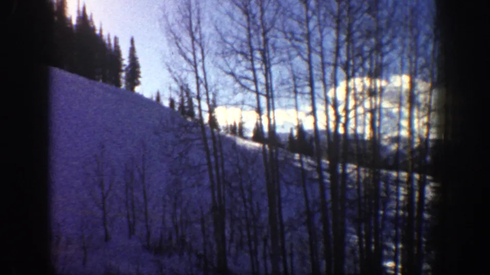 Aspen, Colorado 1961 frame with a steep snow-covered hillside, bare trees, dark evergreens, and bright winter light.