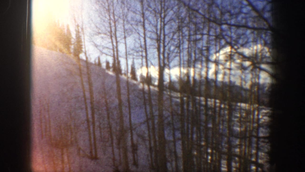 1961 Aspen, Colorado frame with warm sun flare filtering through leafless aspens over a snowy mountain slope.