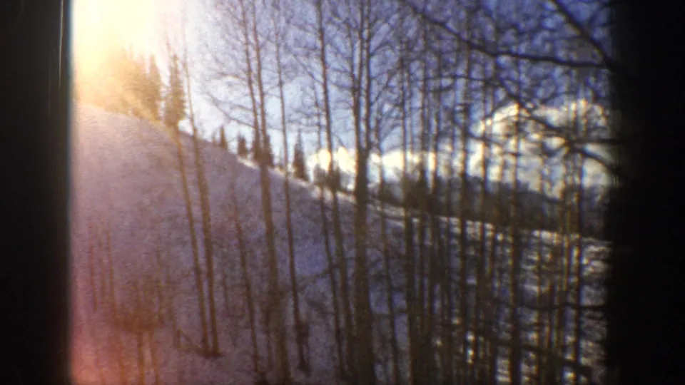 1961 Aspen, Colorado frame with warm sun flare filtering through leafless aspens over a snowy mountain slope.