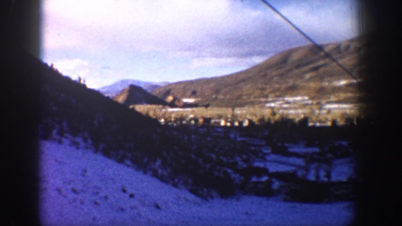 1961 Aspen, Colorado 8mm frame showing snow-covered hills, a shadowed valley settlement, distant mountains, and a diagonal cable near the upper right.