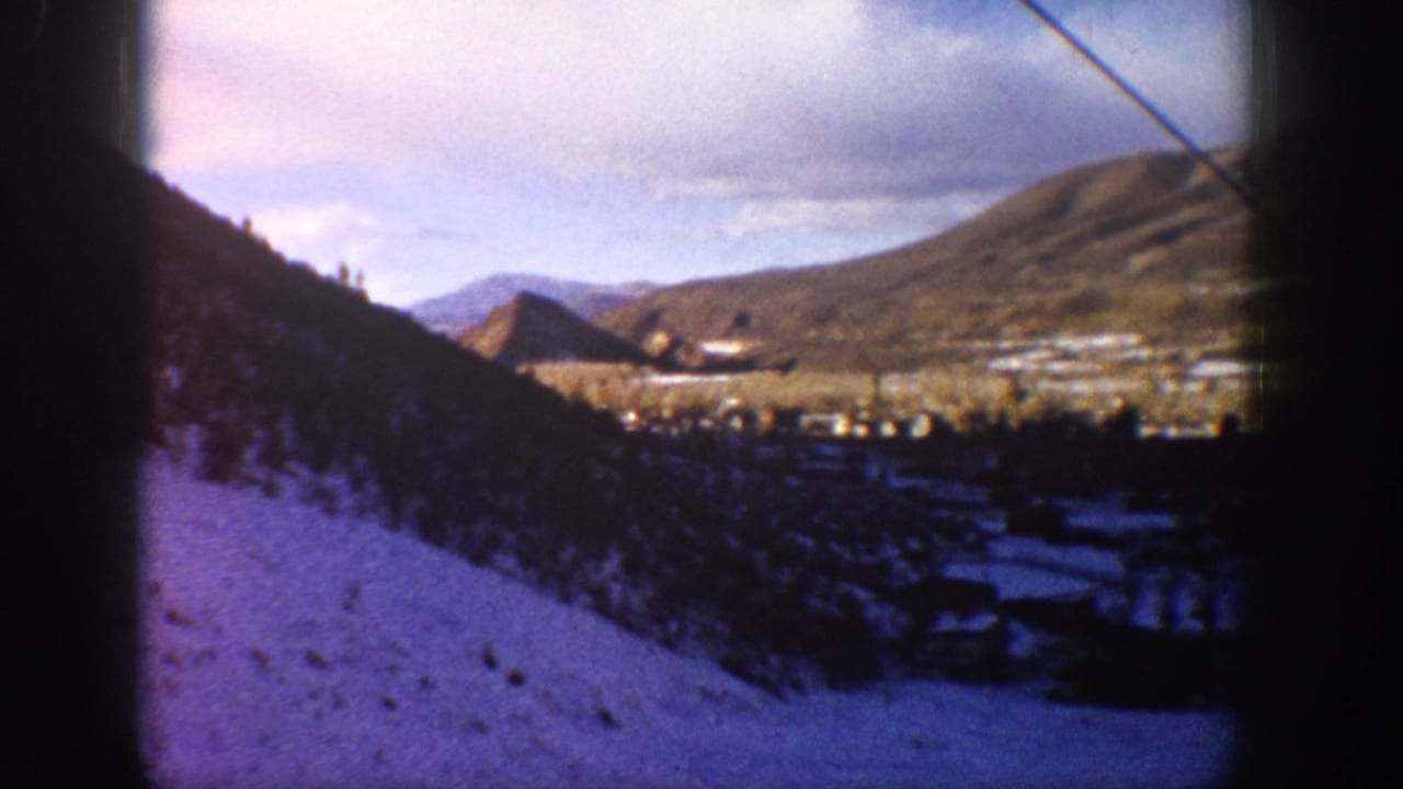 1961 Aspen, Colorado frame with a snowy foreground slope, valley buildings, distant hills, cloudy sky, and visible 8mm frame-edge shadowing.