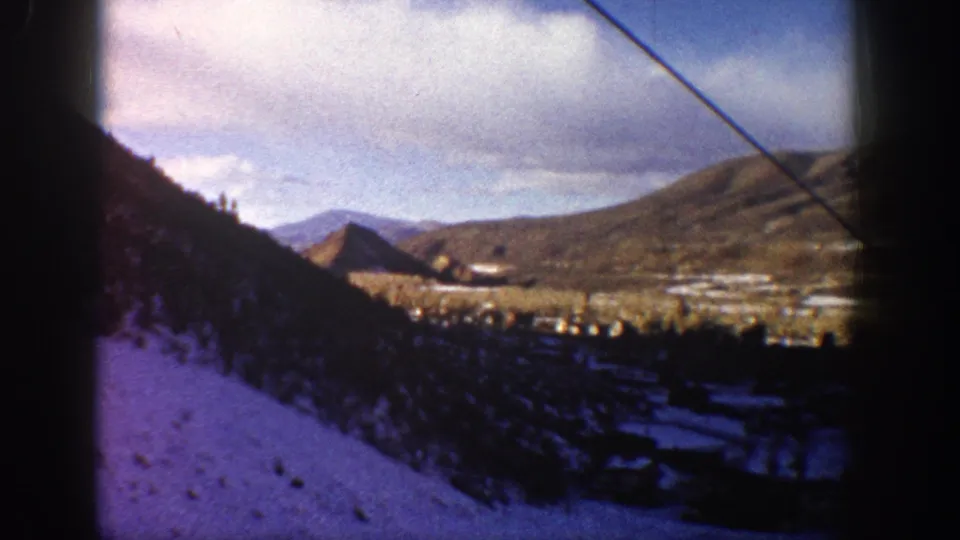 1961 Aspen, Colorado home-movie frame of snowy slopes, a small valley settlement, distant mountains, clouded sky, and pronounced dark film-edge borders.