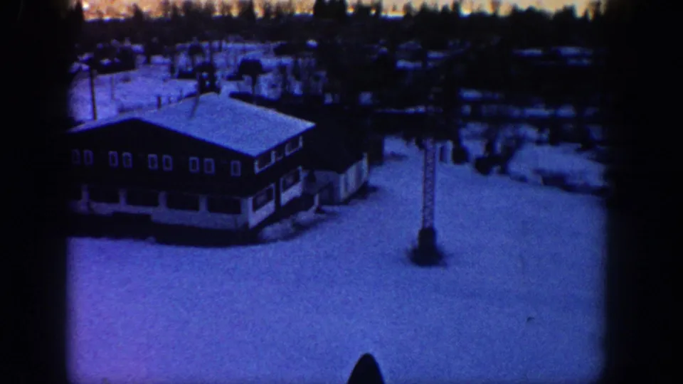 Blue-tinted 8mm frame from 1961 Aspen showing a snowy resort lodge, lift tower, trees, and scattered valley buildings.