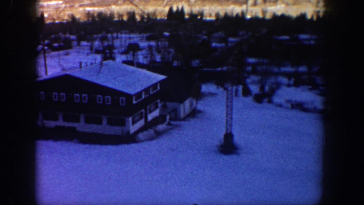 1961 Aspen home-movie frame showing a snow-covered ski lodge from an elevated angle, with dark treeline and a vertical tower nearby.
