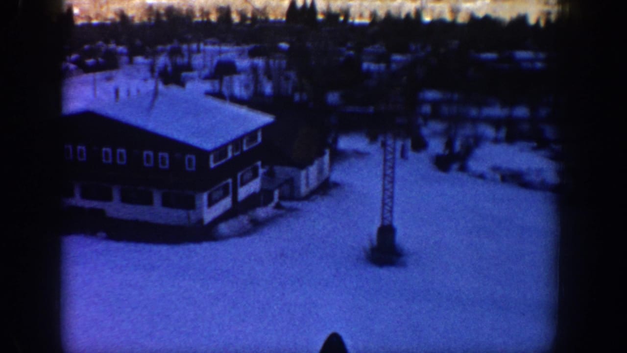Blue-tinted 8mm frame of the Aspen ski lodge area, with snow on the roof, dark trees, and a tower structure in the foreground.